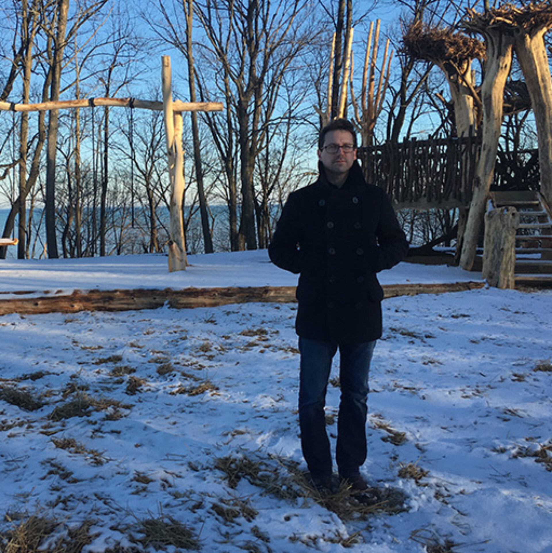 A person stands in snow near wooden structures in a forested area, with a clear blue sky and trees in the background.