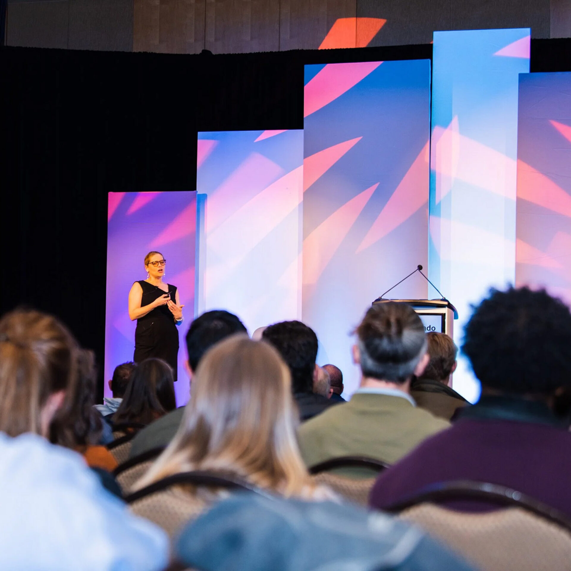 A speaker in a black dress gestures on stage, with colorful light panels behind, while an audience of attentive listeners observes.