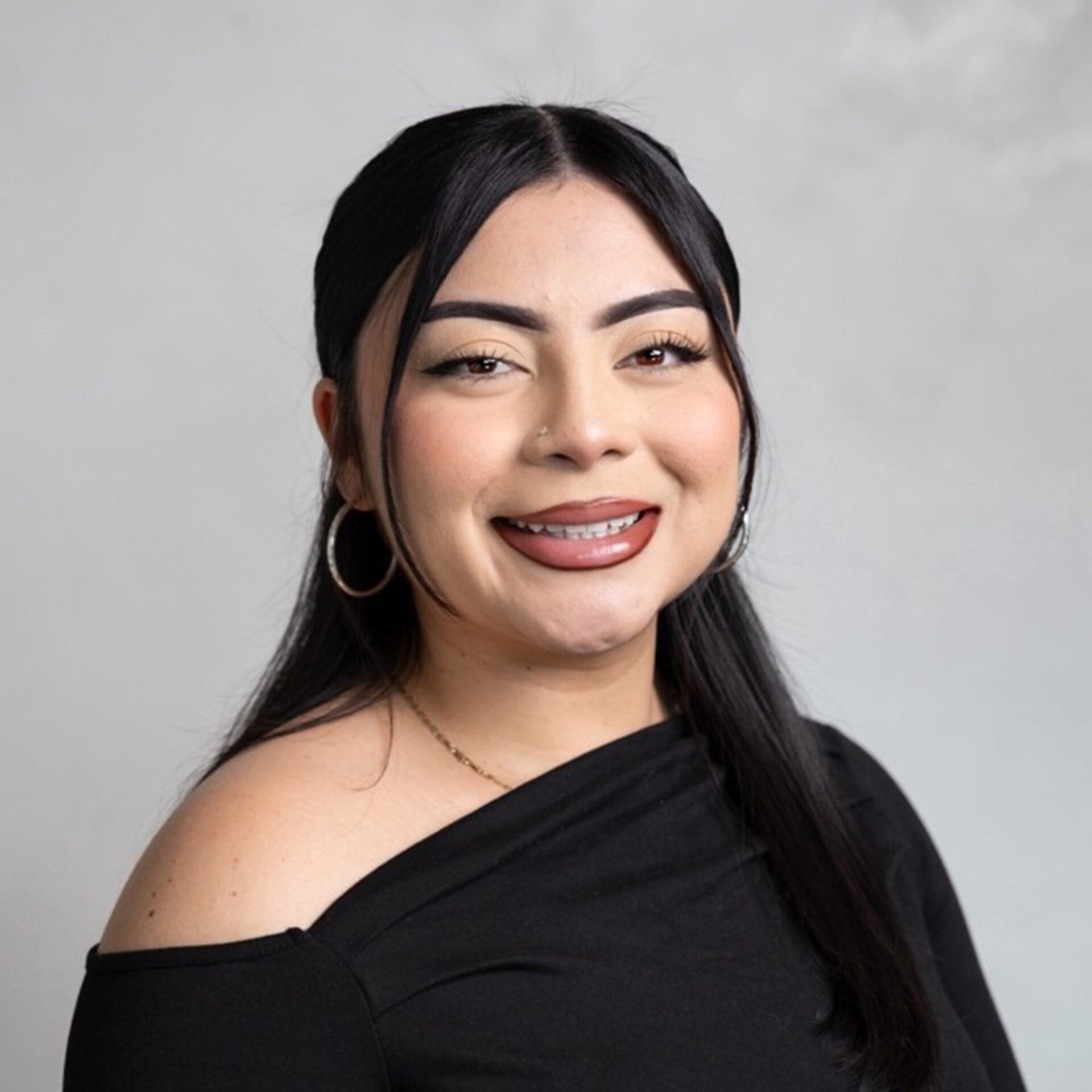 Young woman with long black hair and center part smiles wearing hoop earrings, nose stud, braces, bold brows and an off-shoulder black top.