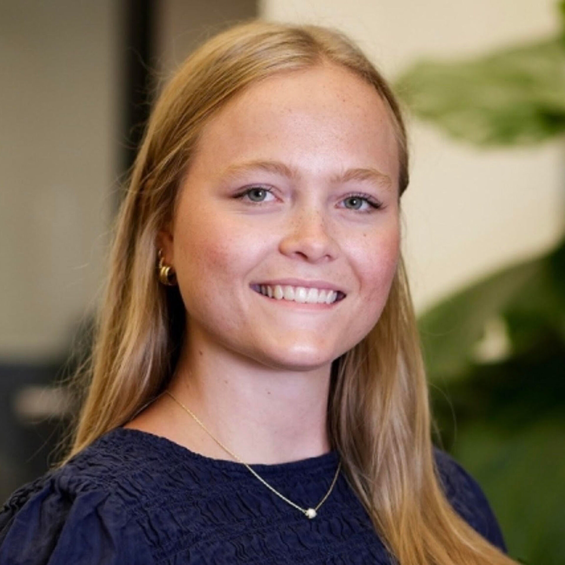 Blonde woman smiling, wearing a navy blouse, gold hoop earrings and a delicate pendant necklace, blurred green plant in the background.