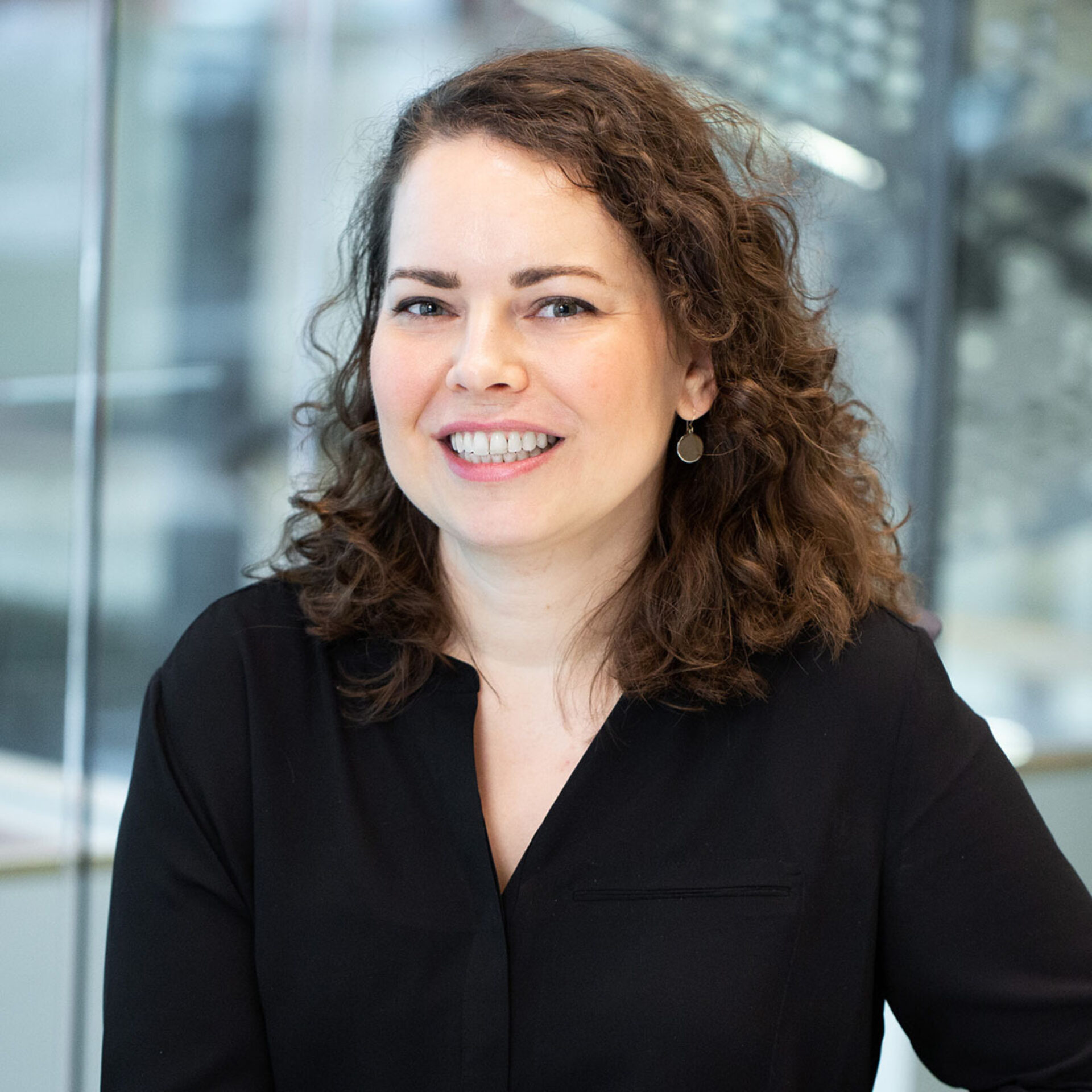 Smiling woman with shoulder-length curly brown hair wearing a black blouse and round drop earrings, posed in a modern glass office setting.