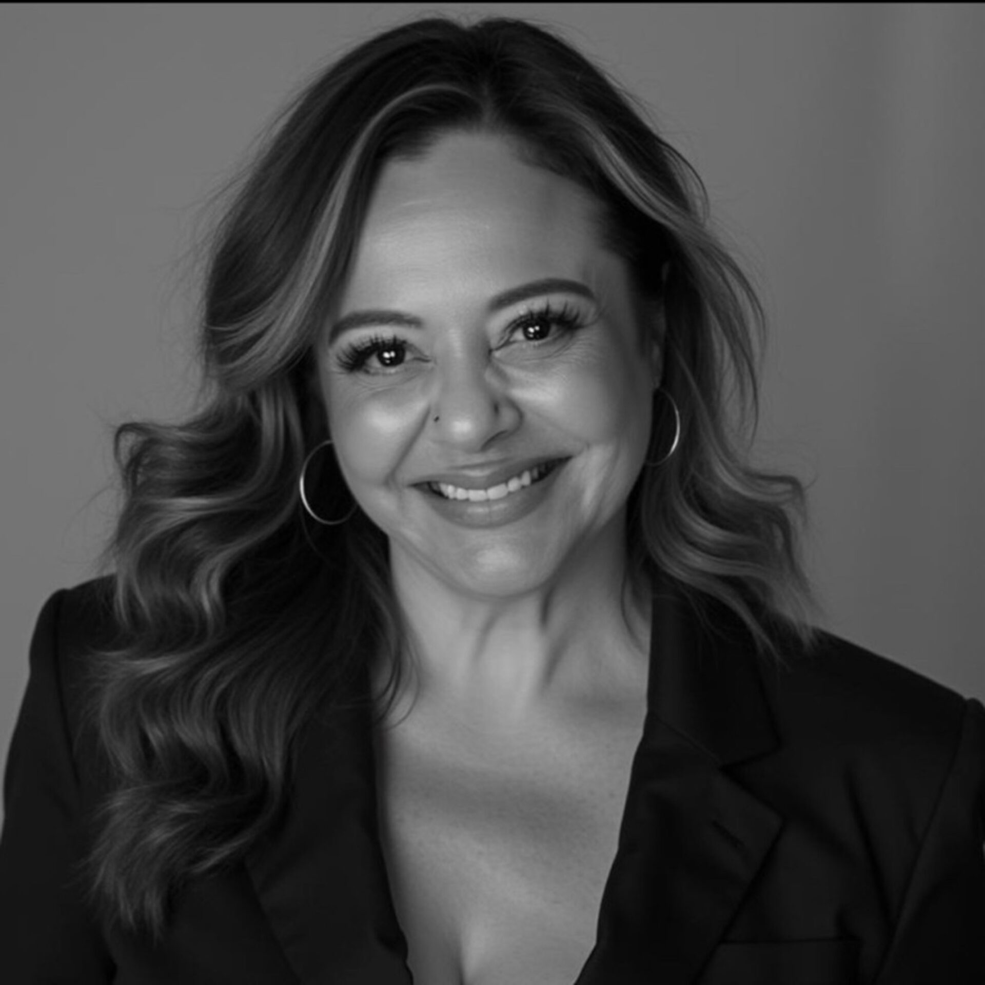 Black-and-white headshot of a smiling woman with wavy shoulder-length hair, hoop earrings, subtle makeup, blazer, and a small nose mole.