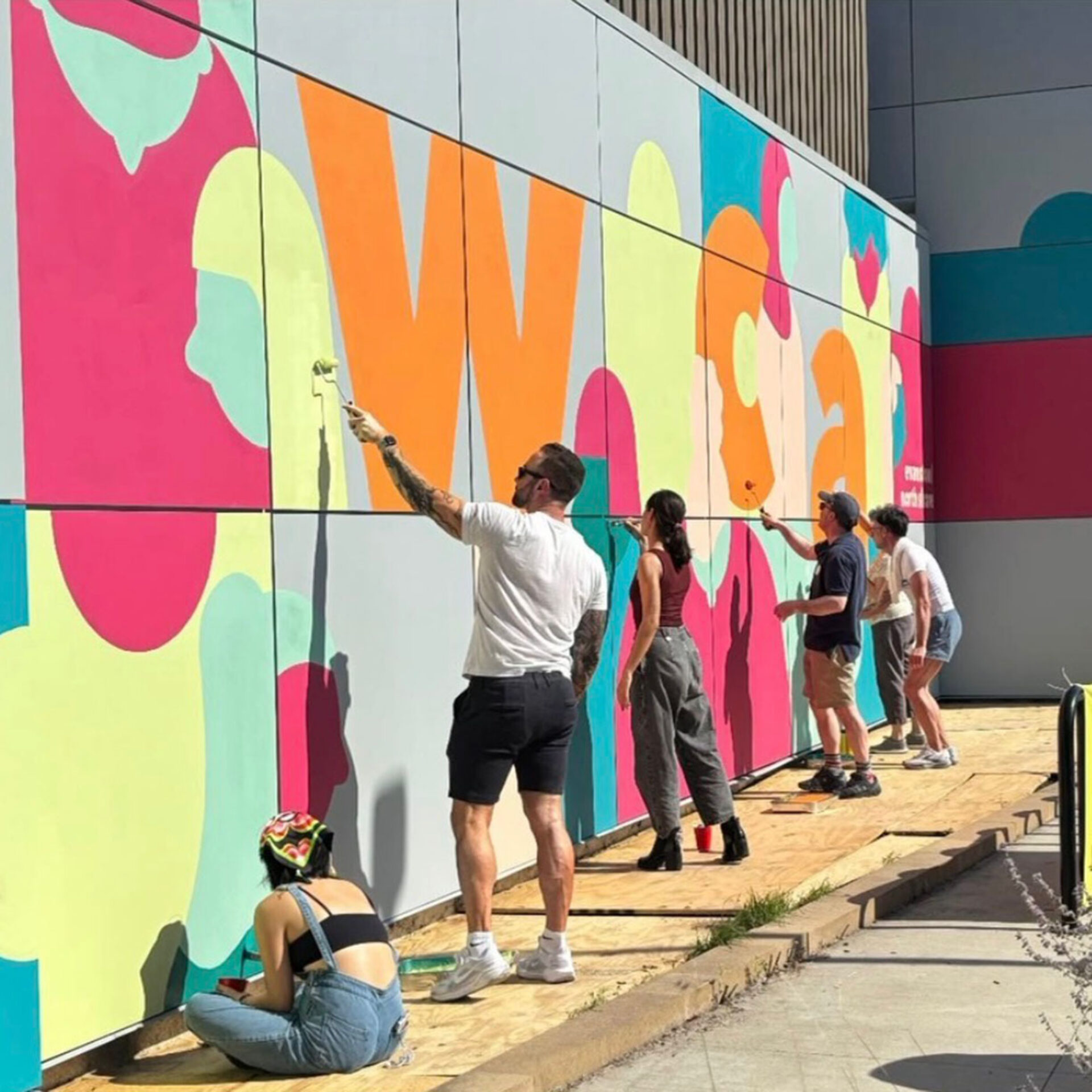 A diverse group of artists paints a vibrant mural on a wall, using various colors and tools, while standing and sitting on wooden platforms.