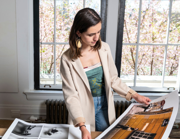 A young woman examines large photographic prints in a bright room, with cherry blossoms visible through the window behind her.