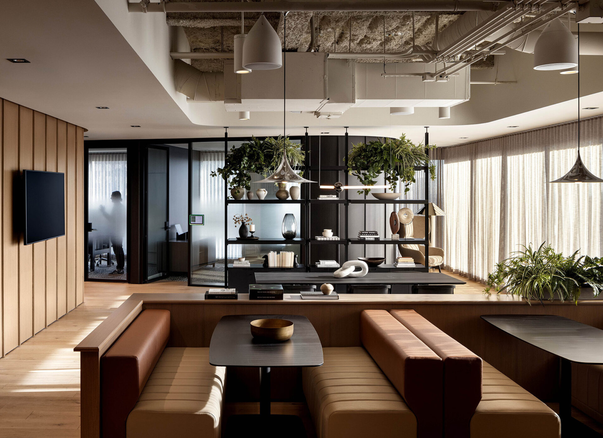 Modern office lounge with leather banquette seating, wood tables, central shelving with hanging plants and frosted glass meeting room.