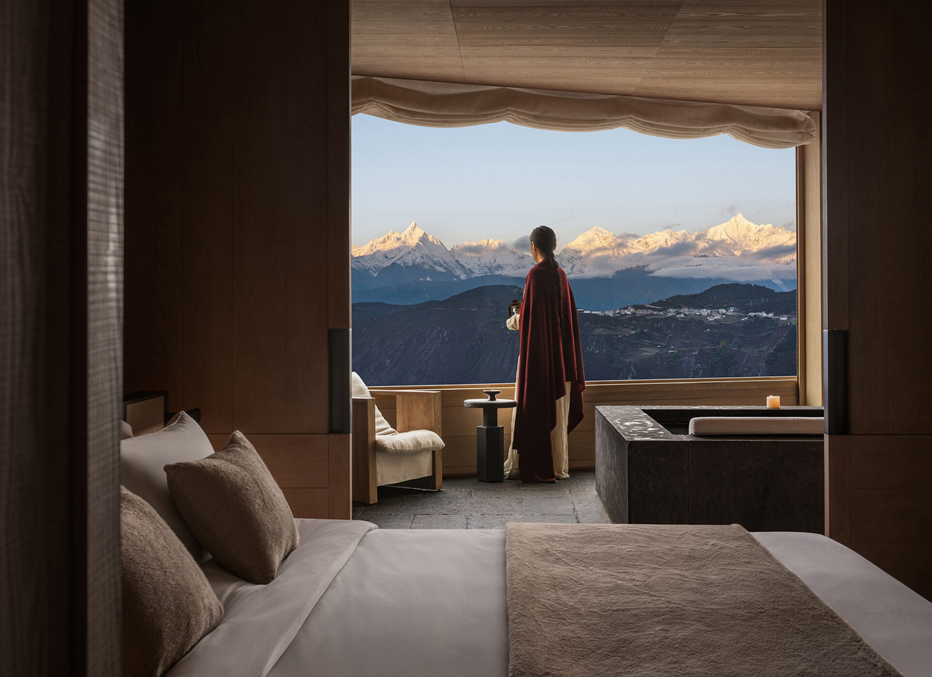 Woman in a red cloak stands at a wide window, holding a cup as sunrise gilds snow-capped peaks above a serene wooden bedroom and stone bath.
