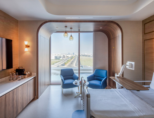 Bright hospital room with a wooden arched window alcove, two blue armchairs, bedside hospital bed with overbed table, city view beyond.
