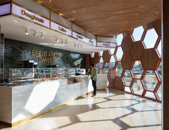 Sunlit modern bakery with marble counter and glass case, hexagonal wooden windows casting honeycomb shadows while a barista serves a patron.