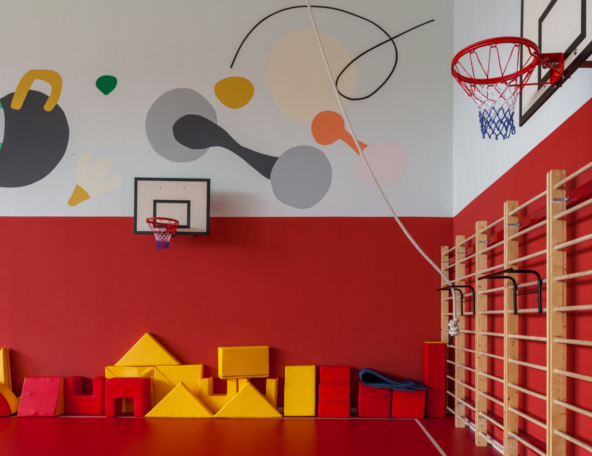 Bright kids gym with red walls, colorful mural, two basketball hoops, hanging climbing rope, wooden stall bars and red/yellow foam shapes.