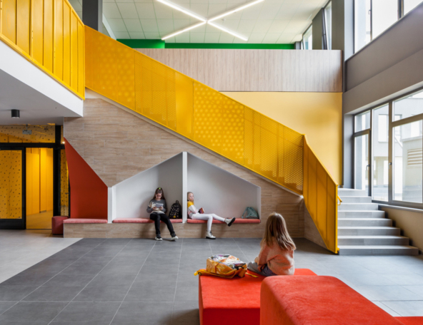 Bright school lobby with yellow diagonal staircase, bench cubbies where two kids read and a third sits on a red bench beside backpacks.