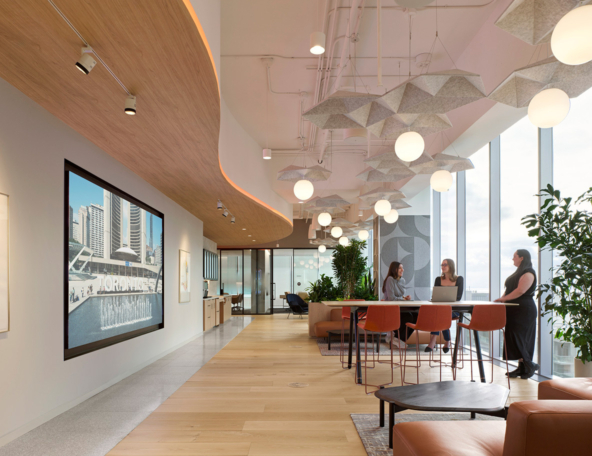 Office lounge with floor-to-ceiling windows, curved wood ceiling, acoustic panels and globe lights above a table where 3 colleagues talk.