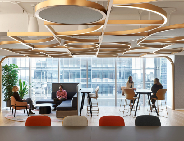 Modern office lounge with circular gold ceiling fixtures, floor-to-ceiling windows, mixed seating groups and people conversing at tables.