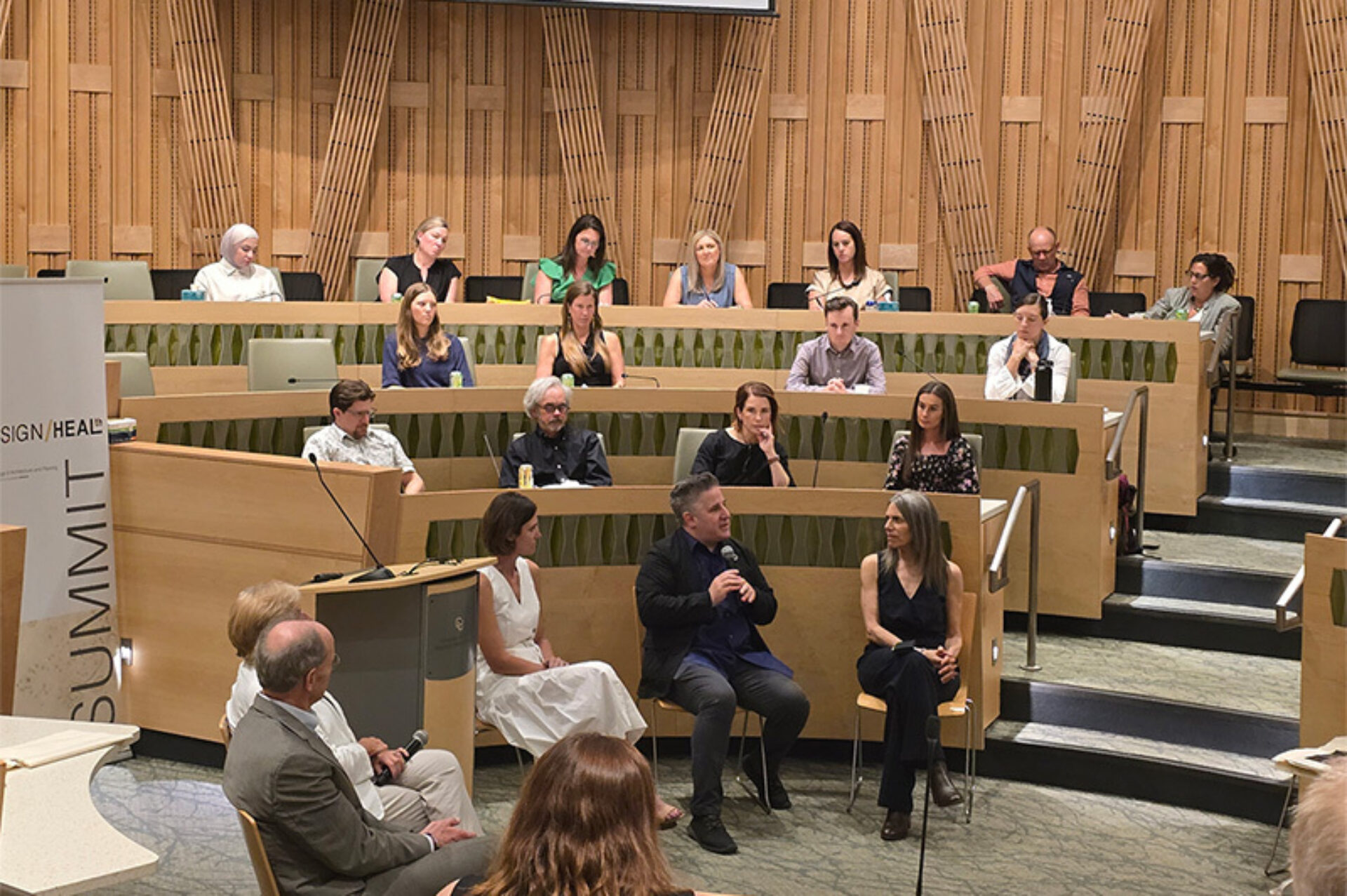Panel discussion in a wood-paneled hall with a seated moderator holding a microphone, two panelists nearby and tiered audience seating.
