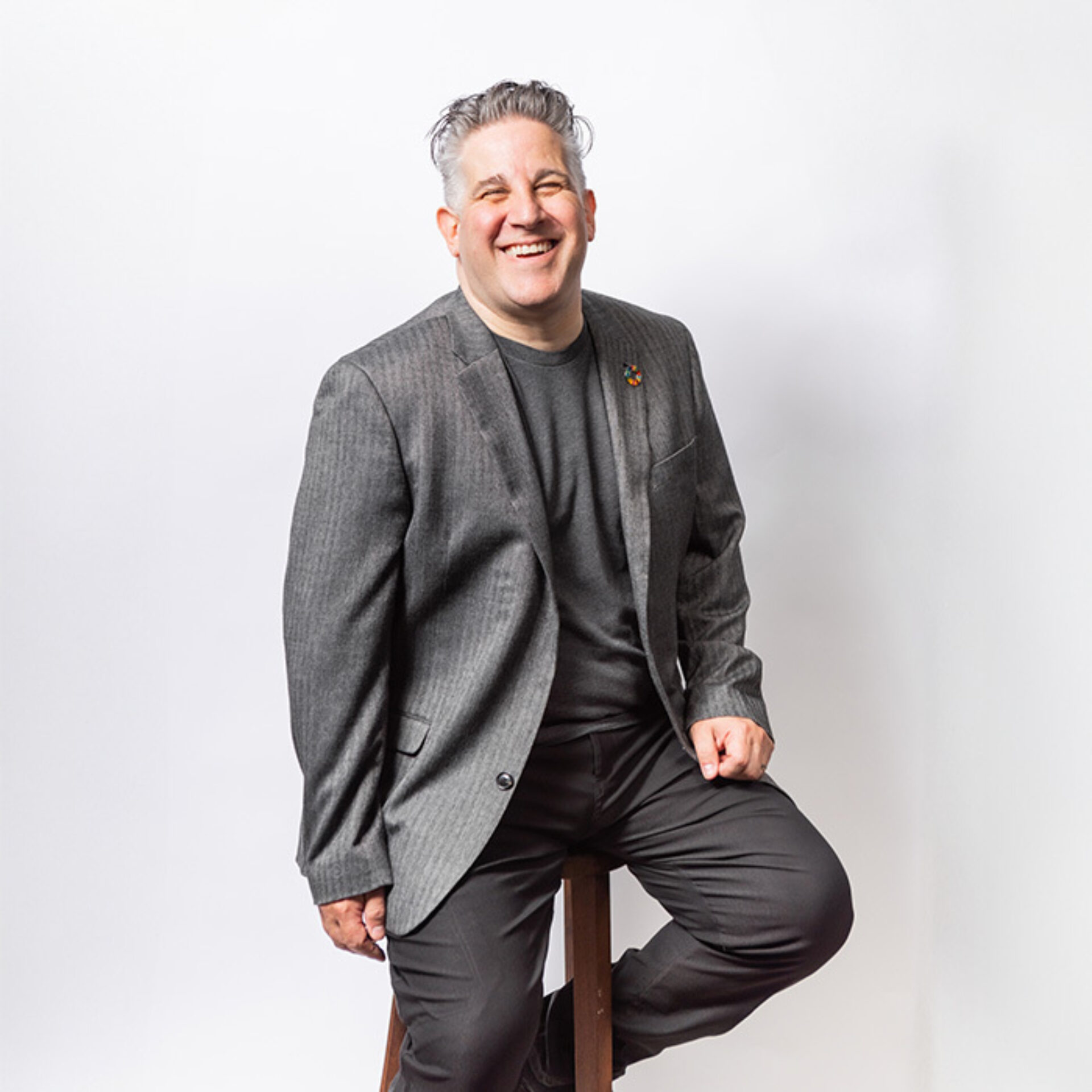 Smiling middle-aged man with salt-and-pepper hair, wearing a grey blazer and black shirt, seated on a wooden stool against white backdrop.