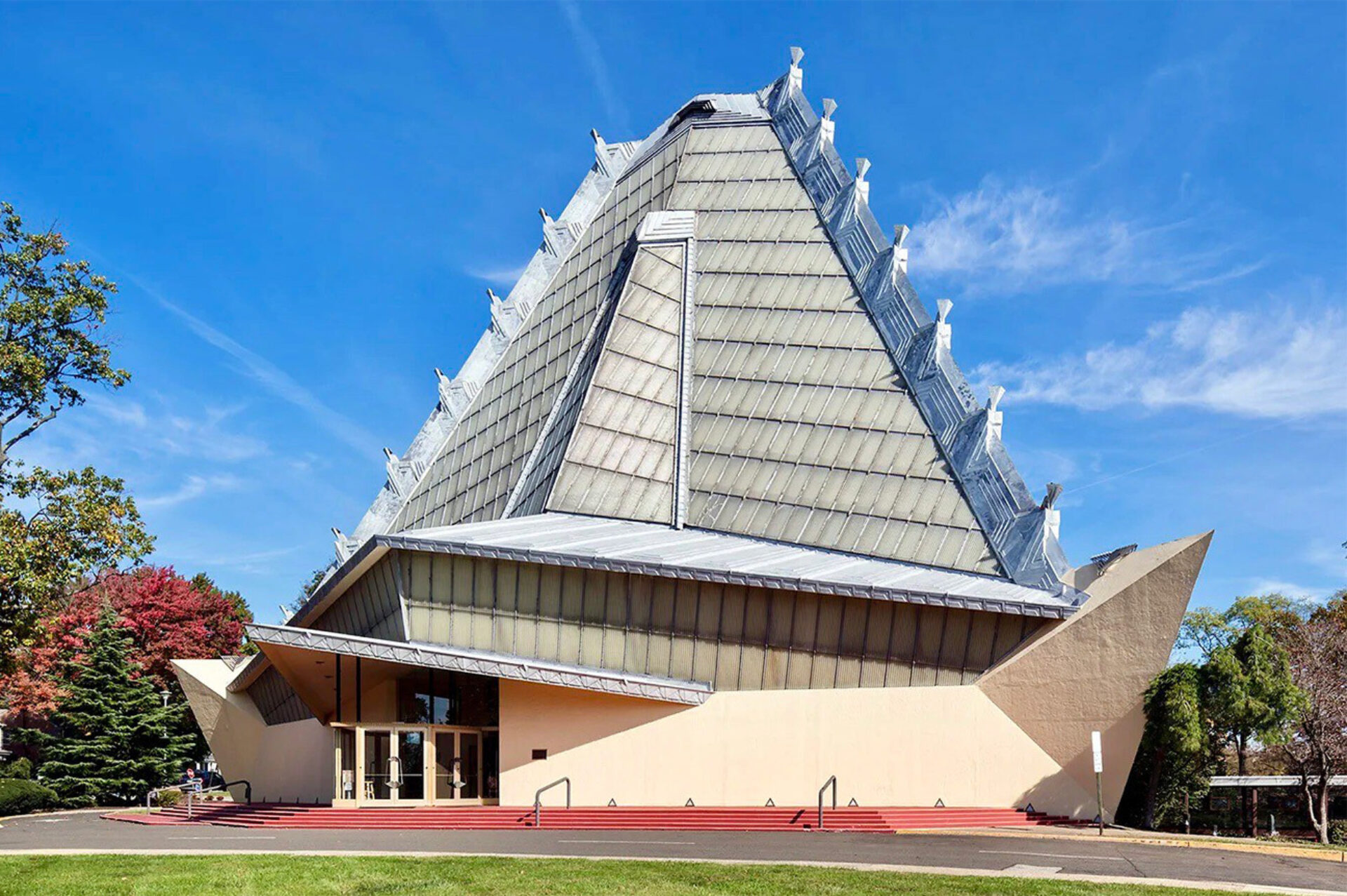 A modern A-frame chapel with steep glass-paneled triangular roof topped by metal finials, beige stucco base, red entry steps and trees.