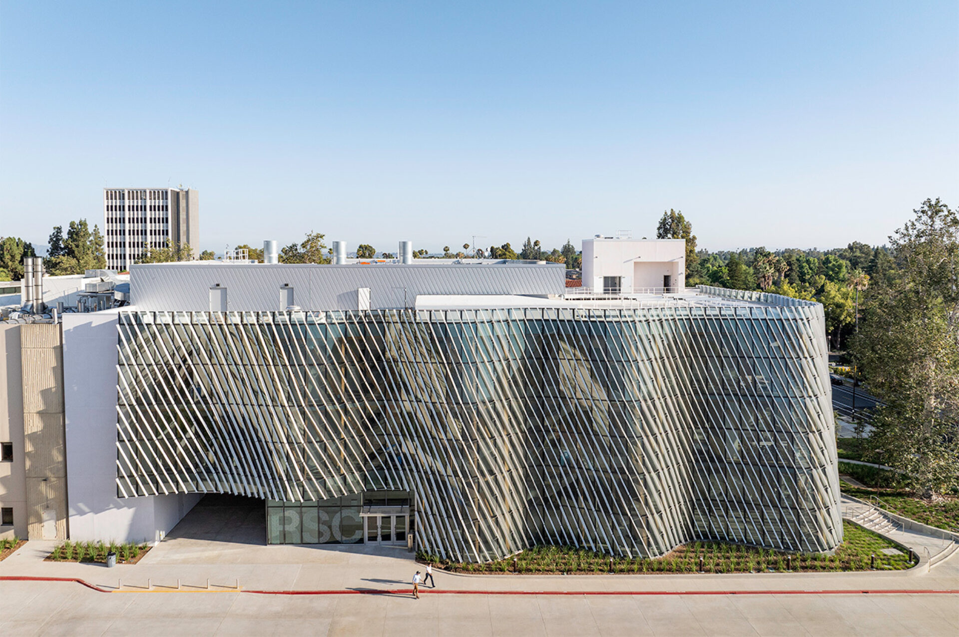 Curved modern building with angled metal fins over a glass facade, two pedestrians walking on the front plaza beneath a clear blue sky.