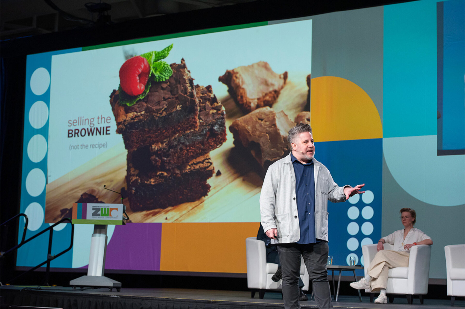 A speaker gestures on stage beside a podium, large slide of stacked chocolate brownies reading 'selling the BROWNIE', panelist seated.