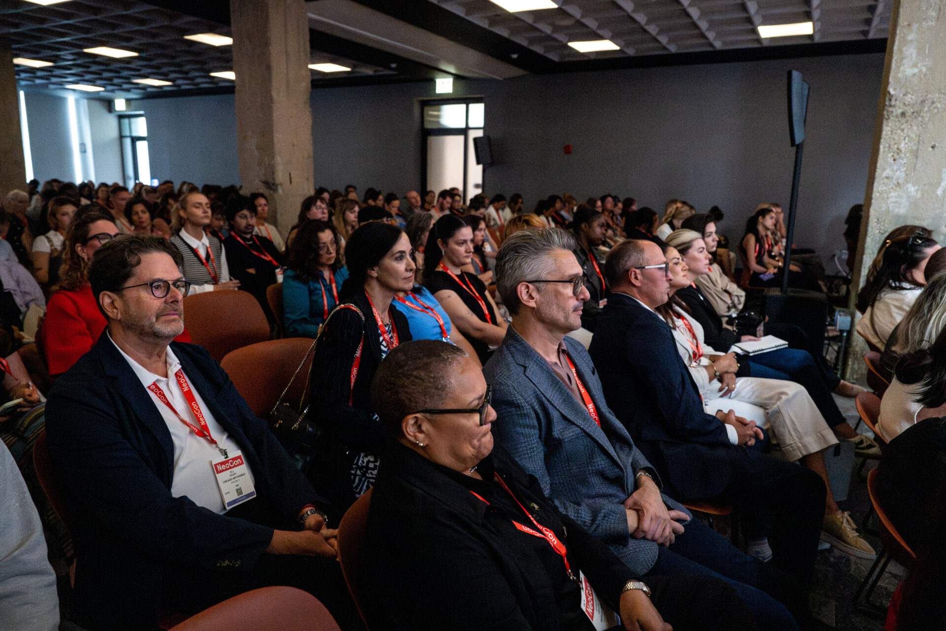 Crowded conference room with diverse attendees wearing red lanyards seated in rows, attentively facing forward and taking notes.