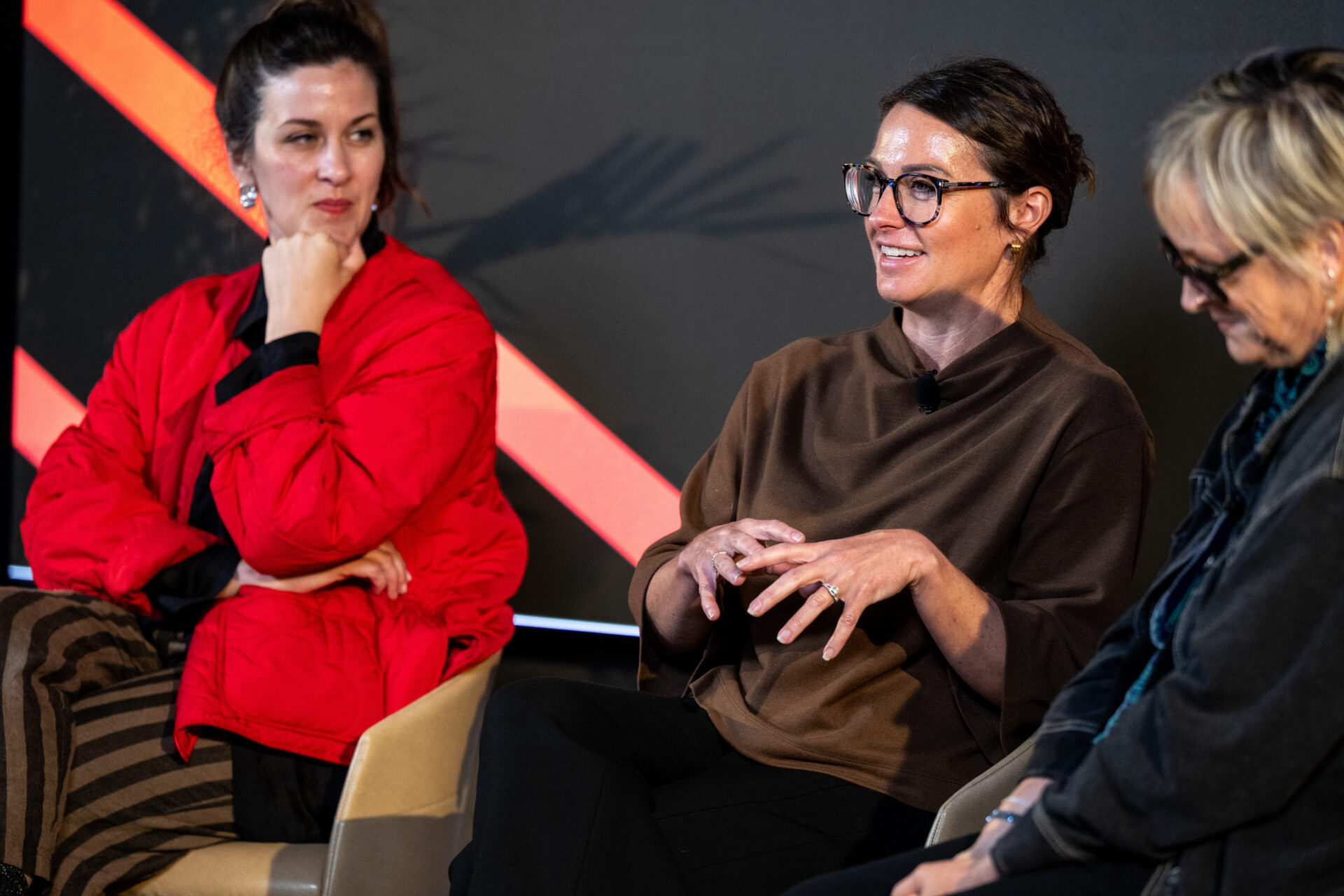 Three women sit on a discussion panel: a smiling woman with glasses gestures, a red-jacketed woman watches, a blonde panelist looks down.
