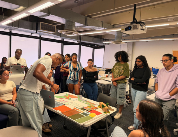 A group of students gather around a table while one student presents colorful architectural models and plans, classmates watching intently.