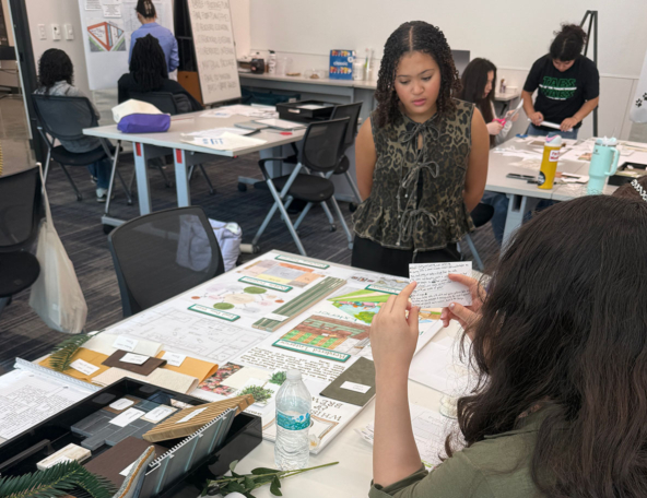 Students in a design studio review presentation boards on a table strewn with swatches, sketches, a water bottle, and note cards.