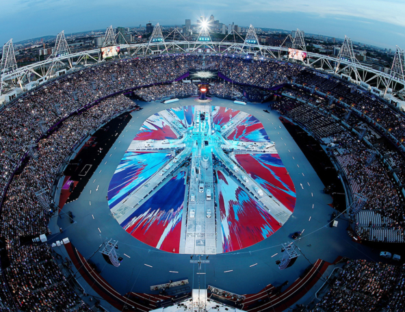 The image captures a vibrant outdoor stadium filled with a large audience, showcasing a colorful performance area with dynamic patterns.