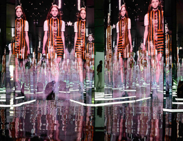 A fashion display features models walking on a mirrored runway, surrounded by colorful screens and reflections in an immersive setting.
