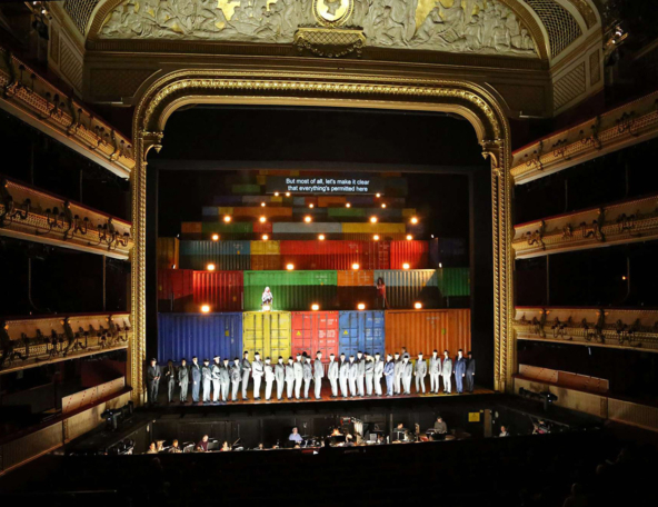 A vibrant stage backdrop features stacked colorful shipping containers, while performers in gray attire stand in formation, highlighting the scene.