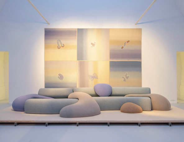 A modern, abstract sofa in soft colors sits against a multi-colored wall tapestry featuring whimsical, embroidered designs.