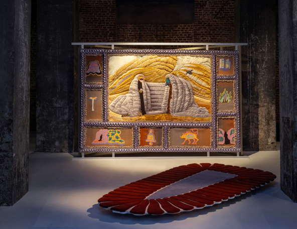 A large textile artwork featuring two figures among hills, surrounded by richly decorated patches, with a unique oval rug below.