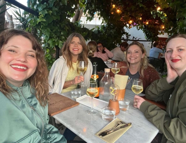 Four smiling women at a marble outdoor table under ivy and string lights, holding white wine glasses with a bottle, menus and water glasses.