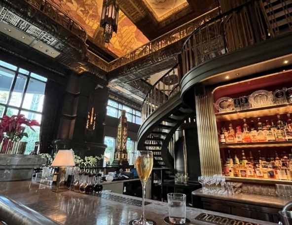 Opulent bar with high ceiling, spiral stairs, backlit liquor shelves and marble counter holding champagne flute, water glass and e-reader.