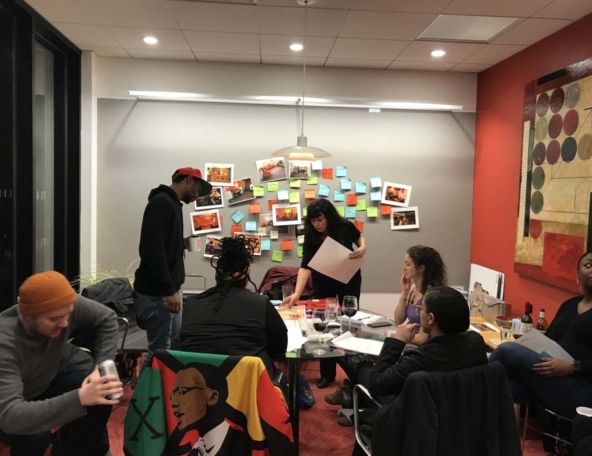 A diverse group gathers around a table in a meeting room; a facilitator pins photos and colorful sticky notes to a gray wall.