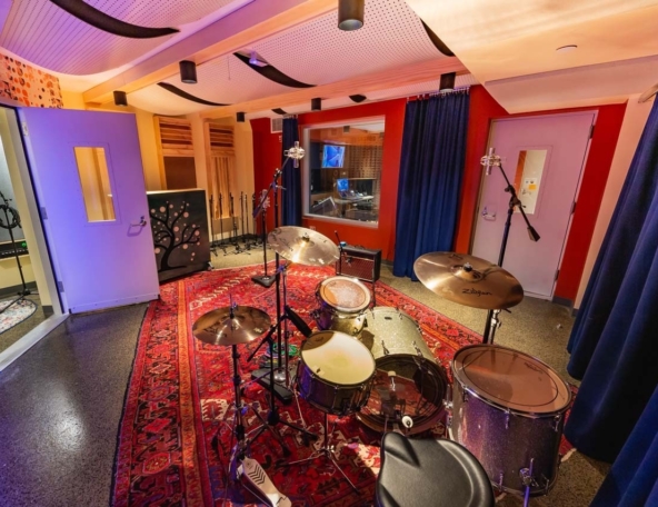 Vibrant recording studio drum room with a silver drum kit on a red Persian rug, overhead microphones, blue curtains, control room window.