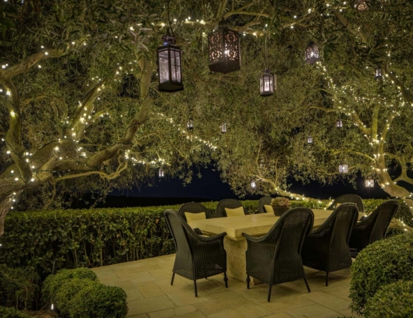 Outdoor stone dining table surrounded by wicker chairs beneath olive trees strung with warm fairy lights and hanging lanterns at night.