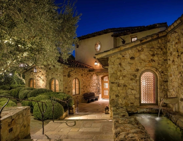 Stone Mediterranean courtyard at dusk with glowing arched windows, lanterns and string lights, manicured boxwood, steps and a small fountain.