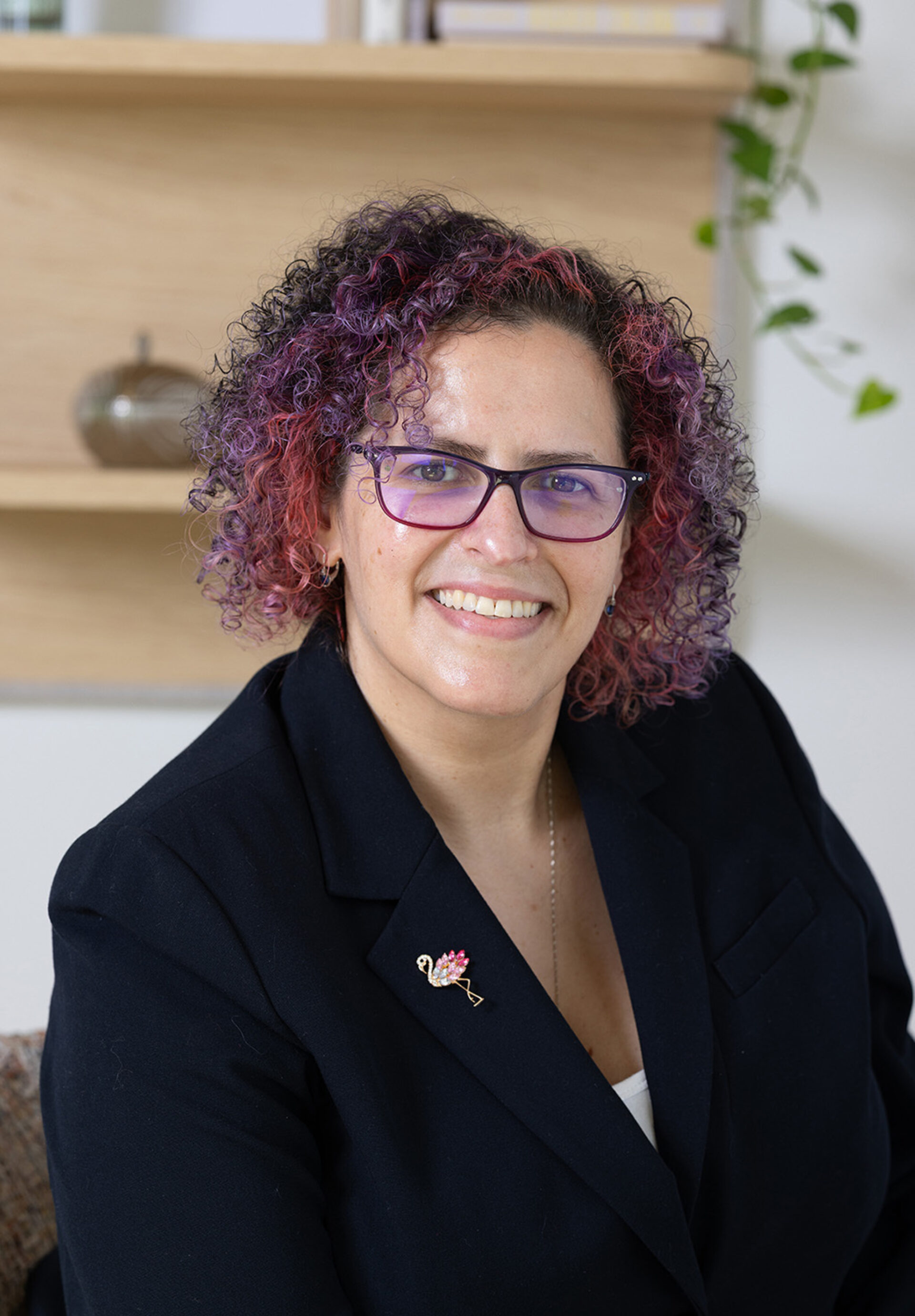 Smiling woman with curly purple-pink hair and glasses wearing a black blazer adorned with a flamingo brooch, bookshelf behind her.