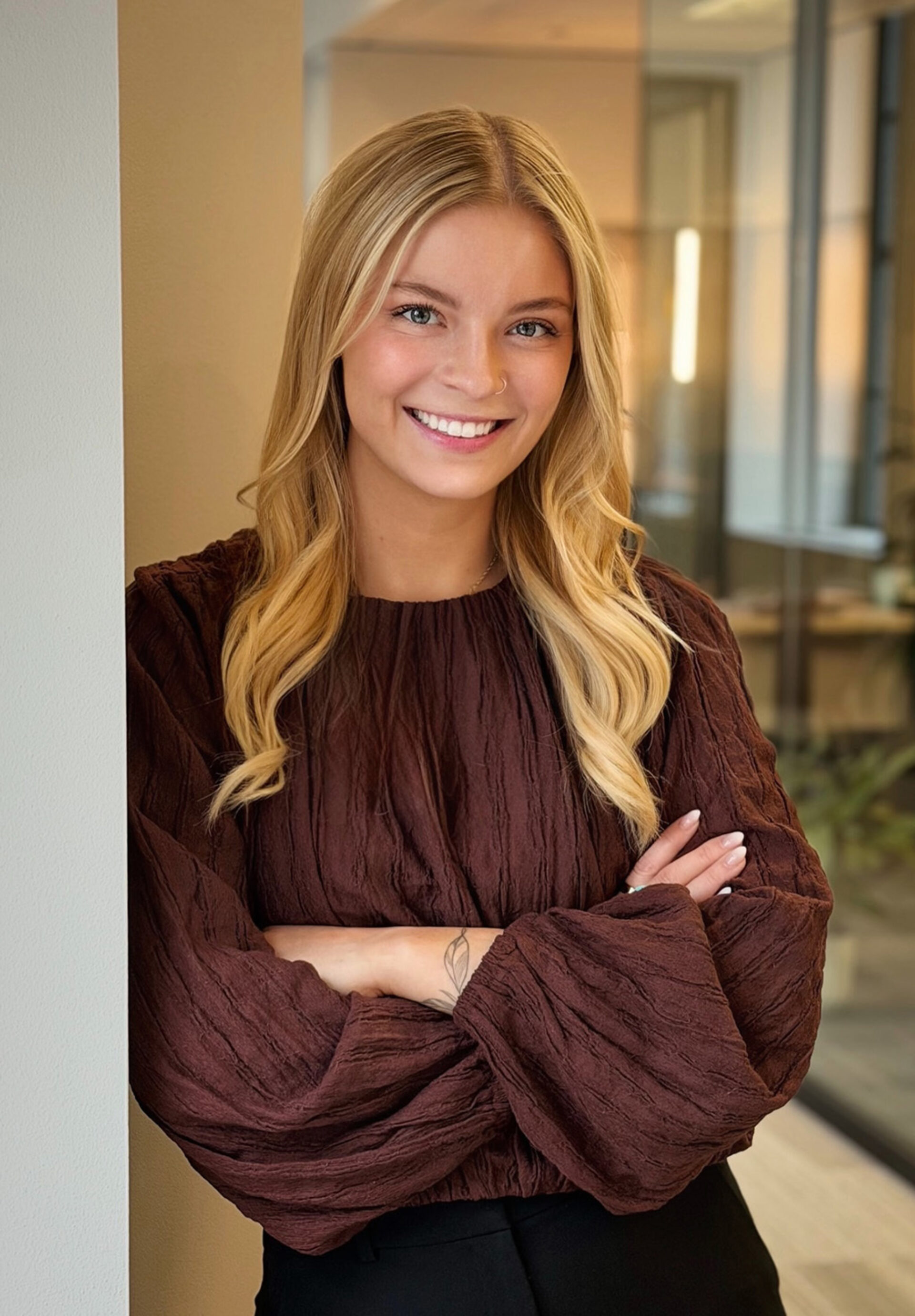 Smiling young blonde with a nose ring, arms crossed, wearing a textured brown blouse with billowy sleeves, standing by an office wall.
