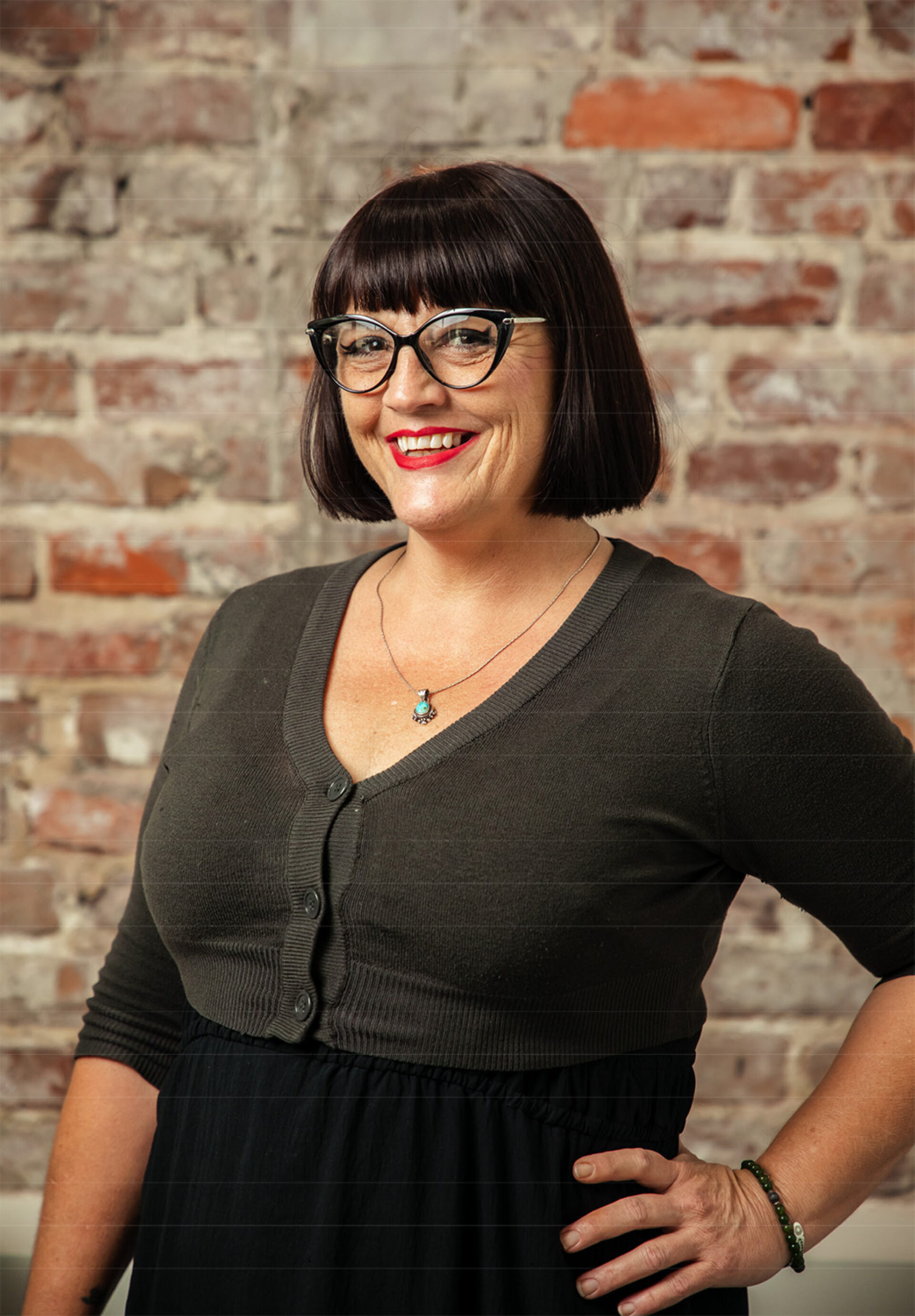 Smiling middle-aged woman with short black bob and bangs, cat-eye glasses, red lipstick, turquoise pendant, green cardigan, hand on hip.