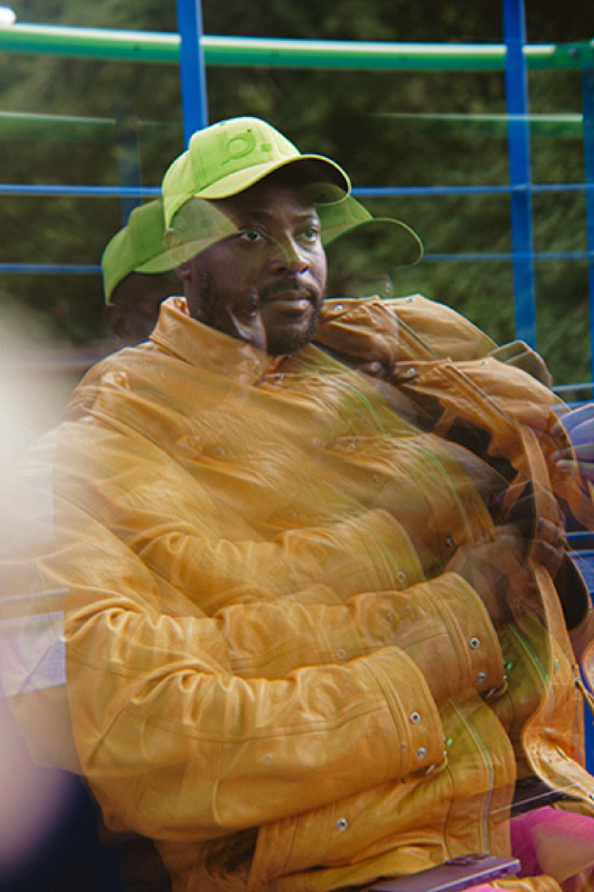 Seated man in a neon green cap and orange puffer jacket sits with arms crossed on colorful outdoor seating, blurred double-exposure effect.