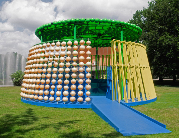 Circular pavilion with green domed roof, hanging rows of beige spheres, blue entry ramp and yellow slatted panels on a grassy lawn.