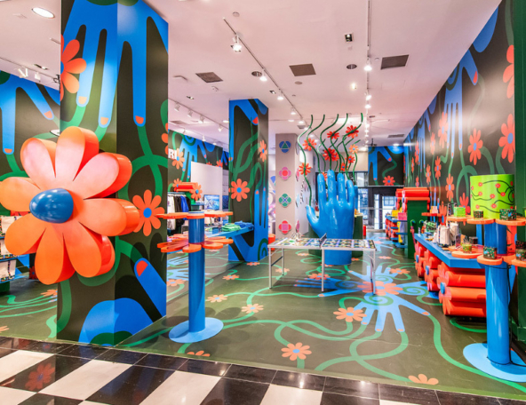 Vibrant boutique interior with oversized blue hands, giant orange flowers, green vine murals on walls and floor, colorful display tables.