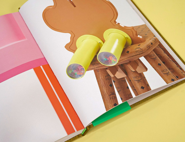 Open colorful book showing wooden chair with yellow binocular-like tubes holding multicolored beads and pink and orange page accents.
