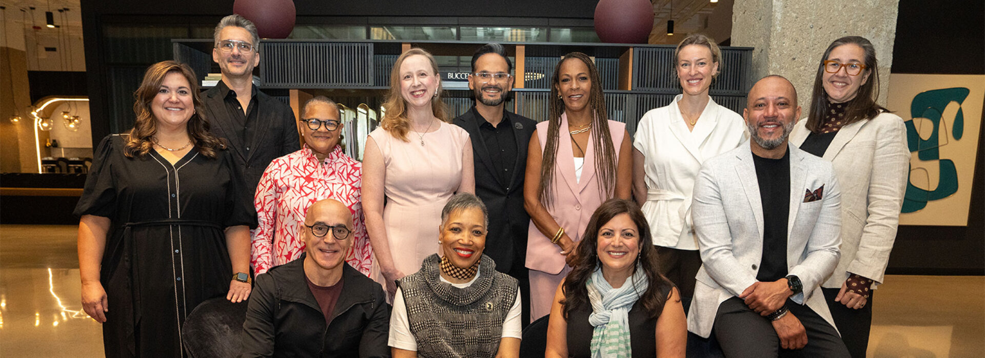 Thirteen diverse smiling professionals, men, women in business and smart casual attire, posed in a modern lobby with art and warm lighting.