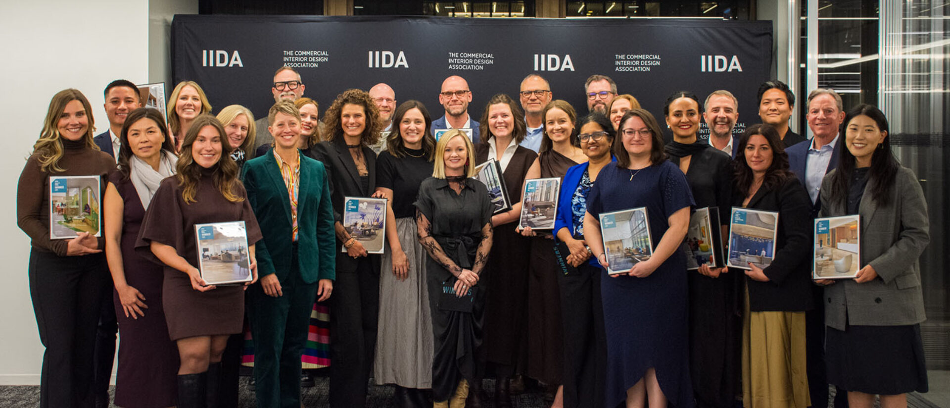 Group of 27 smiling interior designers holding portfolios and awards in front of a black IIDA backdrop at a formal award event.