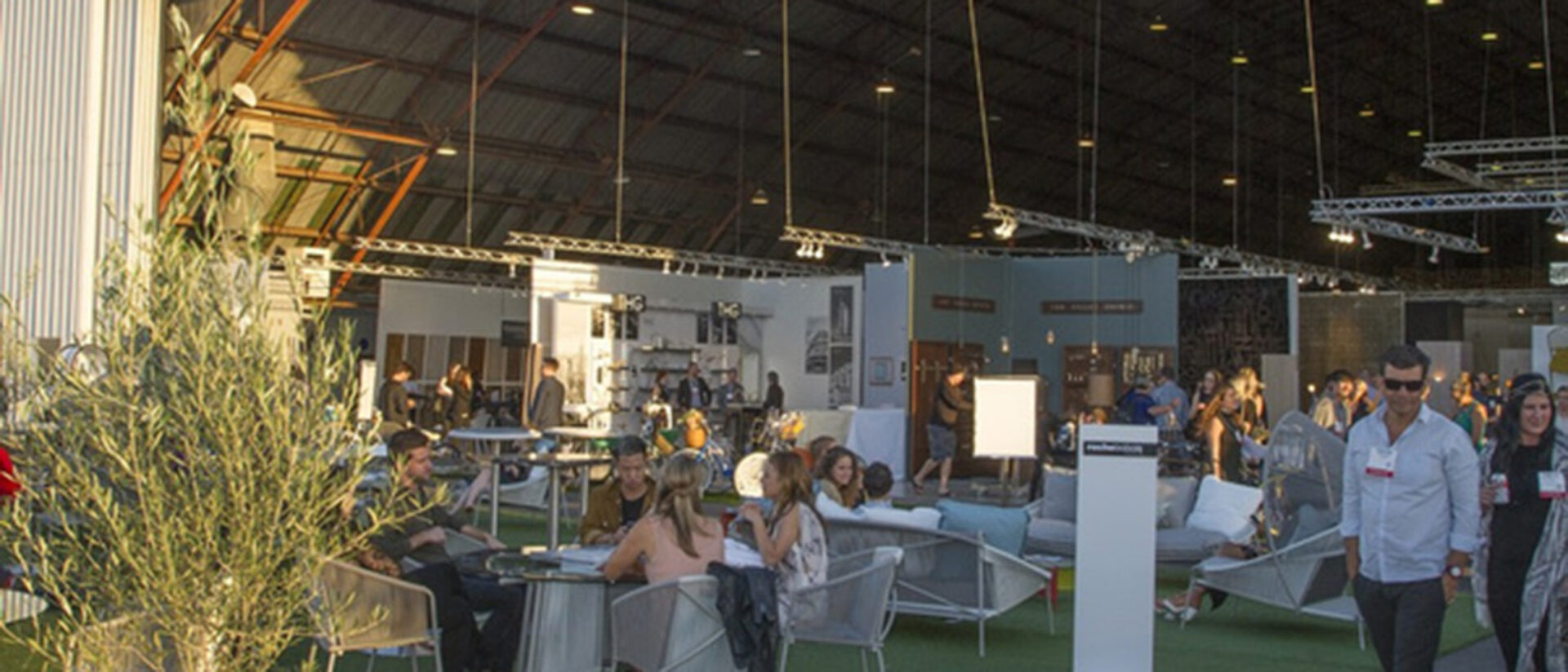 Busy indoor expo in a hangar-like space with overhead trusses and lights, turf flooring, lounge seating with groups talking and demo stage.