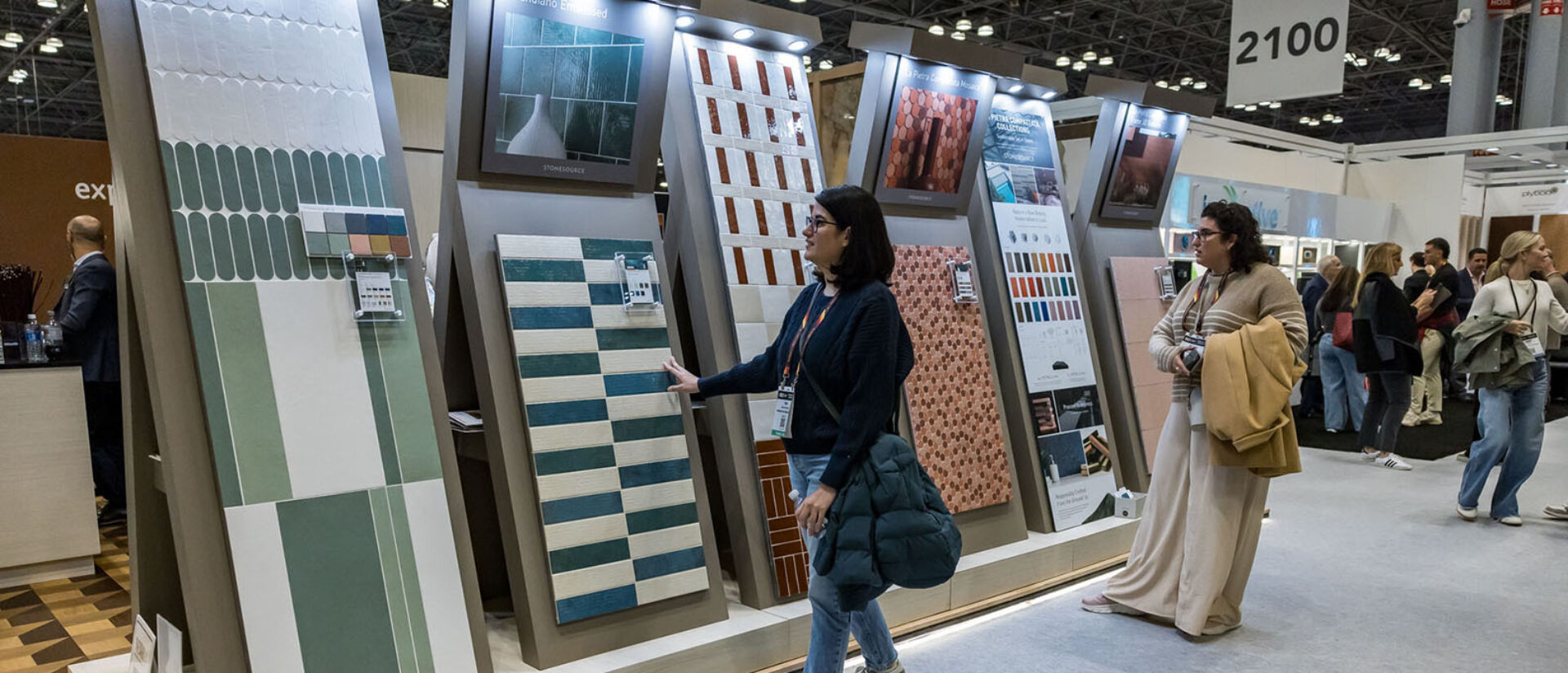 Two women examine large vertical tile display panels at a busy trade show booth, touching striped and hexagonal ceramic tile samples.