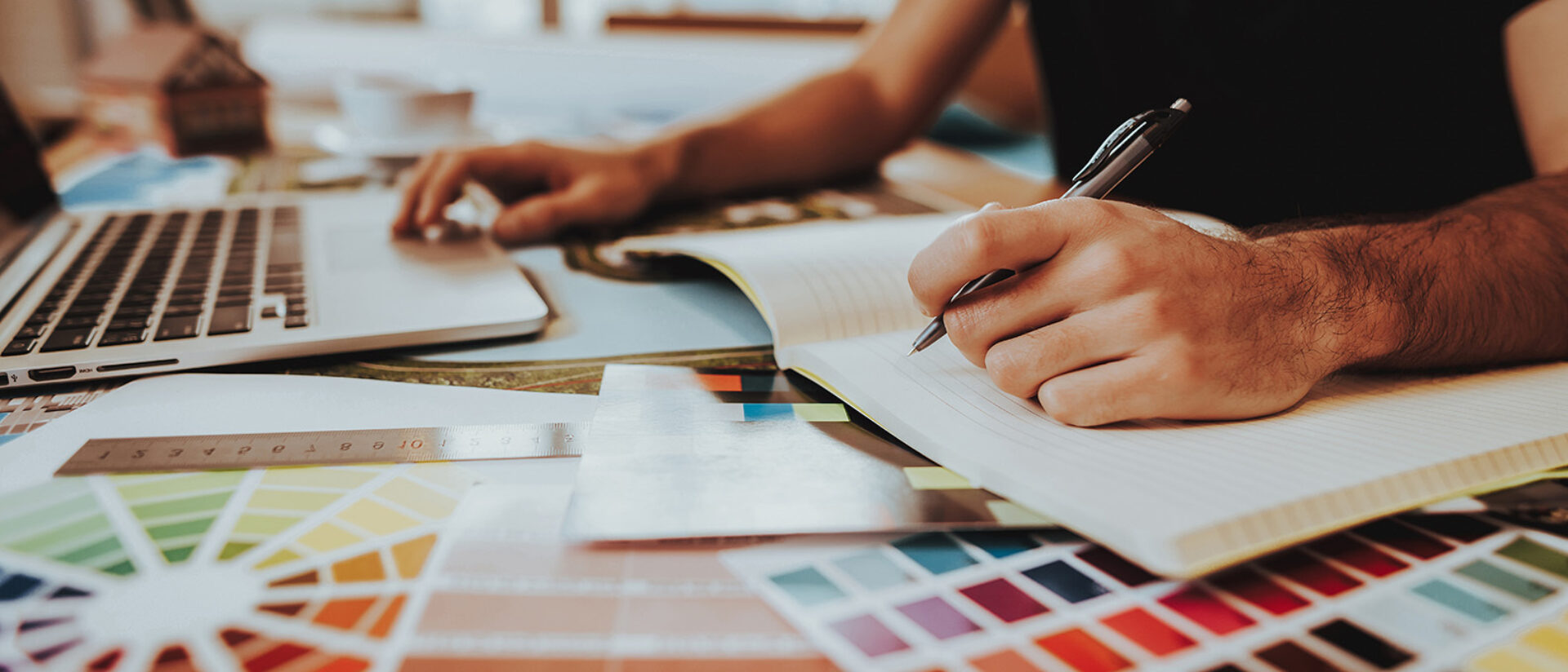 A person is working at a desk with a laptop, writing notes in a notepad, surrounded by color swatches and design tools.