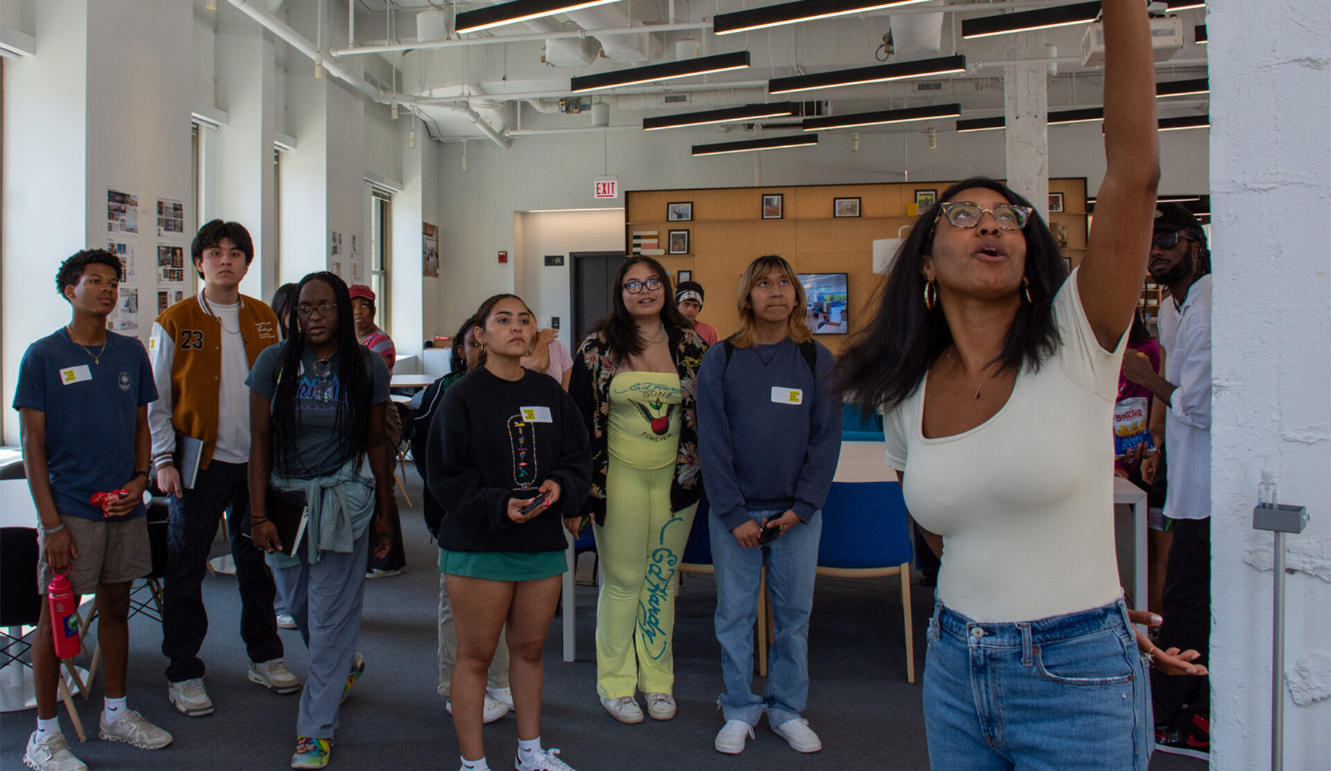 A diverse group of young people attentively watches a woman speaking, gesturing with her arm raised in a bright, modern space.
