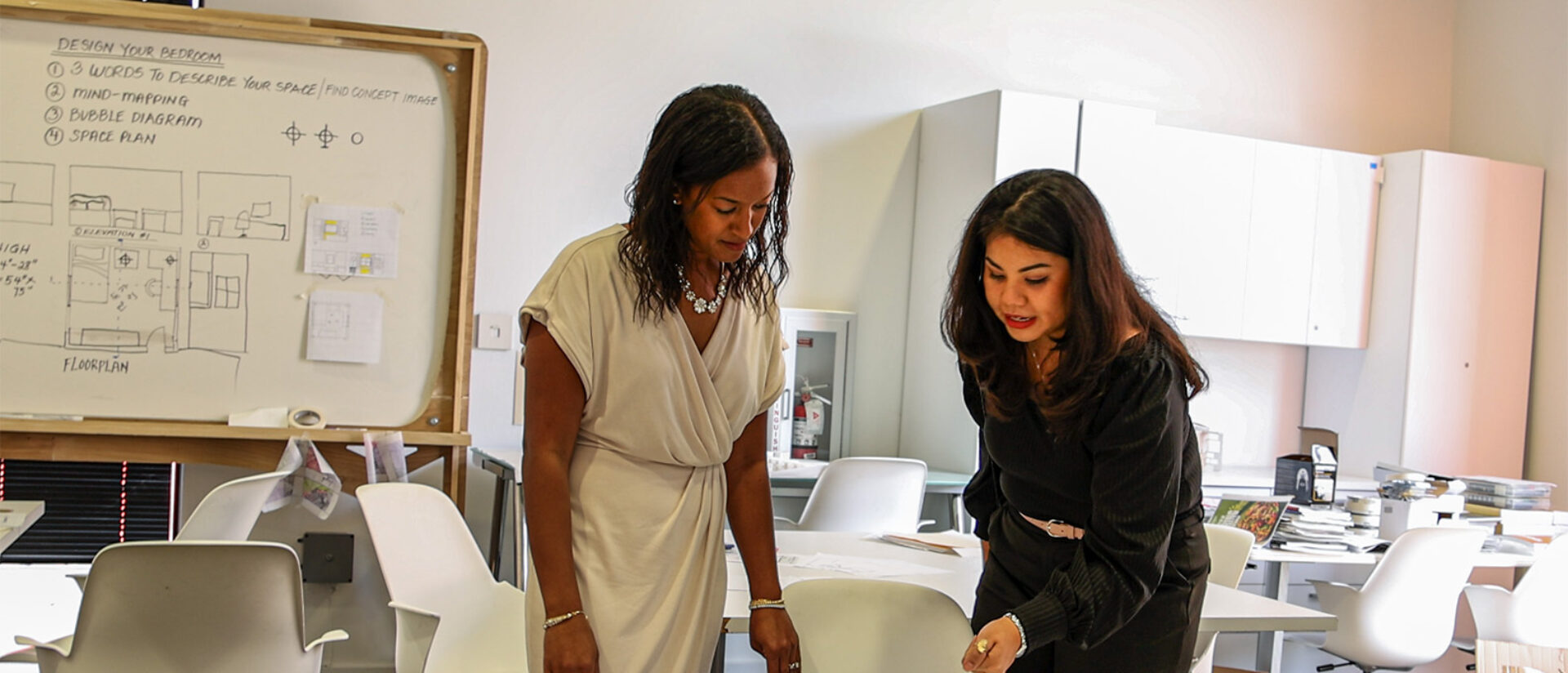 Two women engage in a discussion while reviewing a layout on a table, with a whiteboard featuring design concepts in the background.